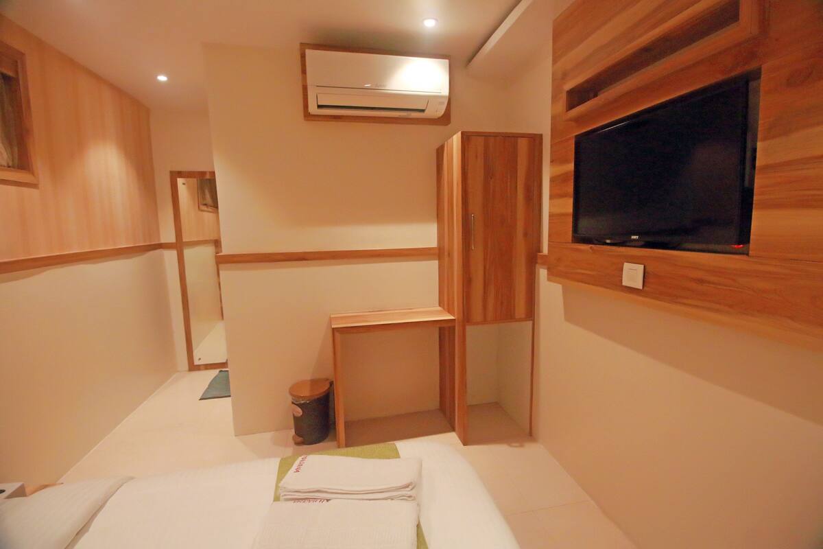 A hotel room with a wooden vanity, a flat-screen TV mounted on the wall, a small wooden table, and a trash can.