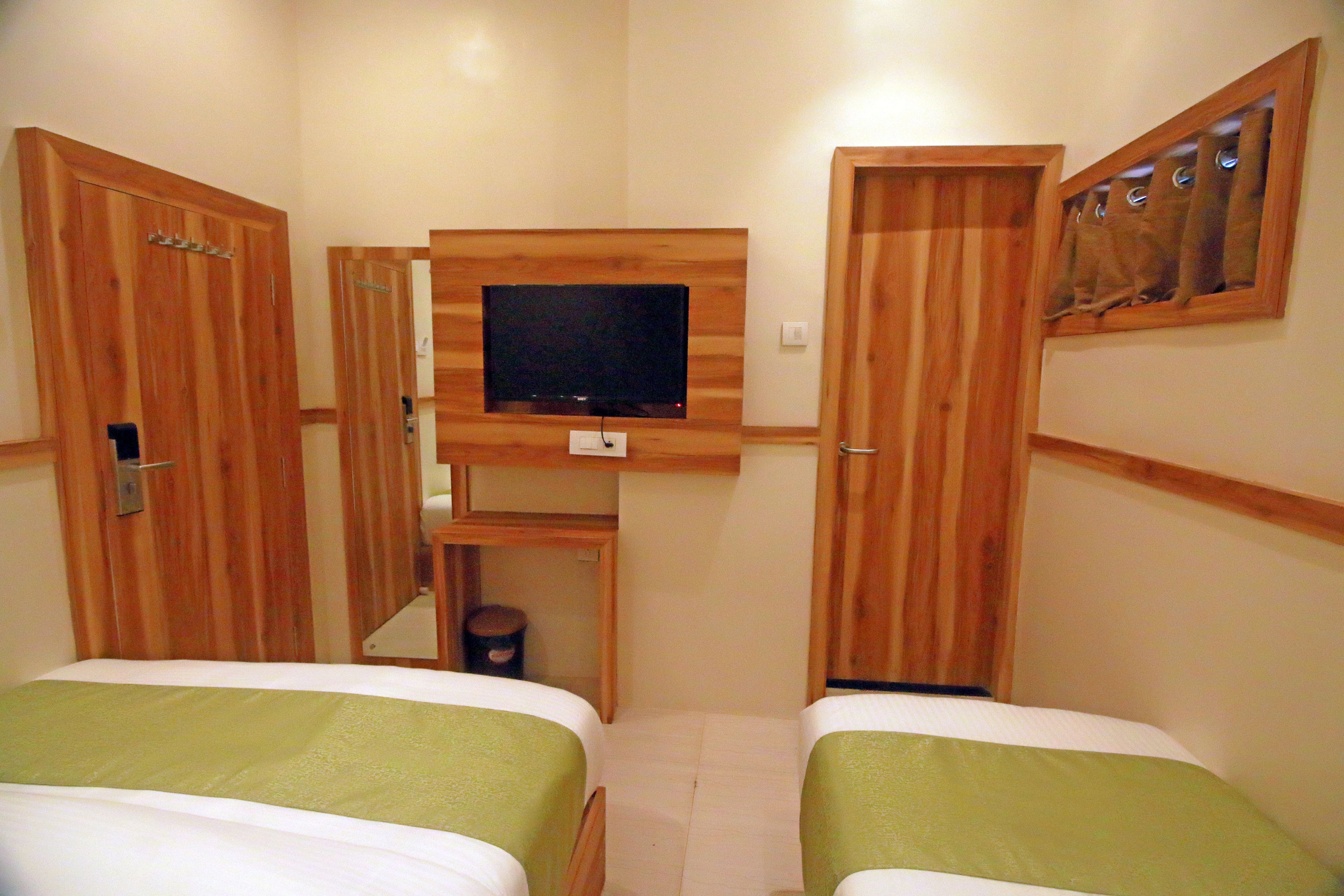 A small room with two beds, a television mounted on the wall, wooden doors, and a small trash bin.