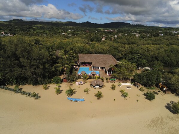 A beachside resort with a swimming pool, lounge chairs, and a thatched-roof building.