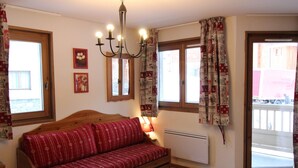 A cozy room with a wooden sofa, a small table, and a chandelier. There are two windows with curtains, a radiator, and a door leading outside. - Spacious apartment in Lanslevillard with balcony and parking (Lanslevillard)