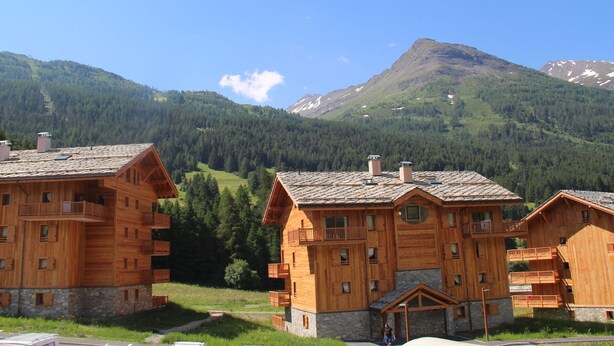 A wooden chalet-style building with multiple balconies and a stone foundation, surrounded by a mountainous landscape. - Spacious apartment in Lanslevillard with balcony and parking (Lanslevillard)