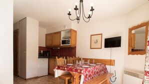 Un coin repas avec une table en bois dressée pour quatre personnes, un lustre, un téléviseur fixé au mur et un four à micro-ondes dans la cuisine.