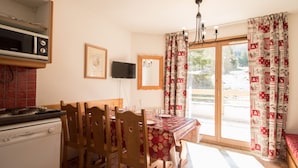 Un coin repas avec une table dressée pour quatre personnes, des chaises en bois, une nappe rouge, un téléviseur fixé au mur et une vue sur l’extérieur grâce à une porte vitrée.