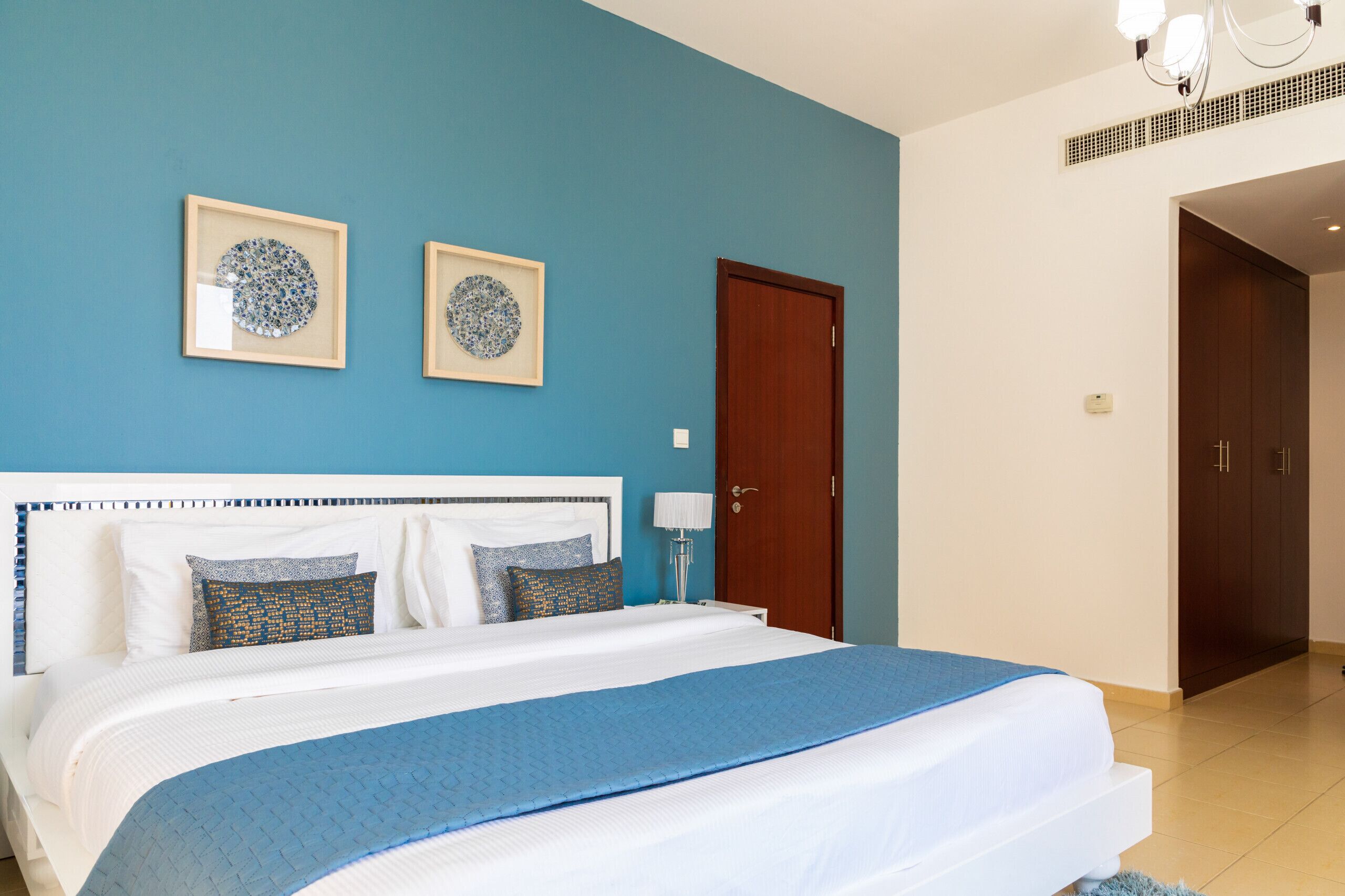A bedroom with a bed, two framed artworks, a wooden door, and a nightstand with a lamp.