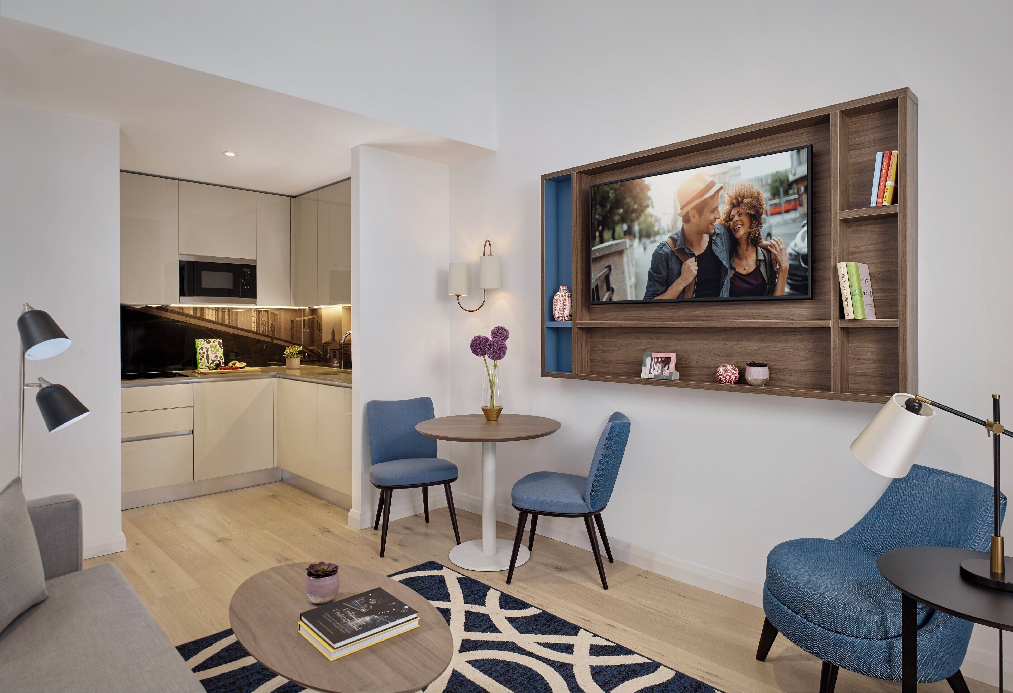 Un soggiorno moderno con un divano, un tavolino da caffè e due poltrone blu. In parete, un televisore montato sopra una mensola in legno con libri e oggetti decorativi.