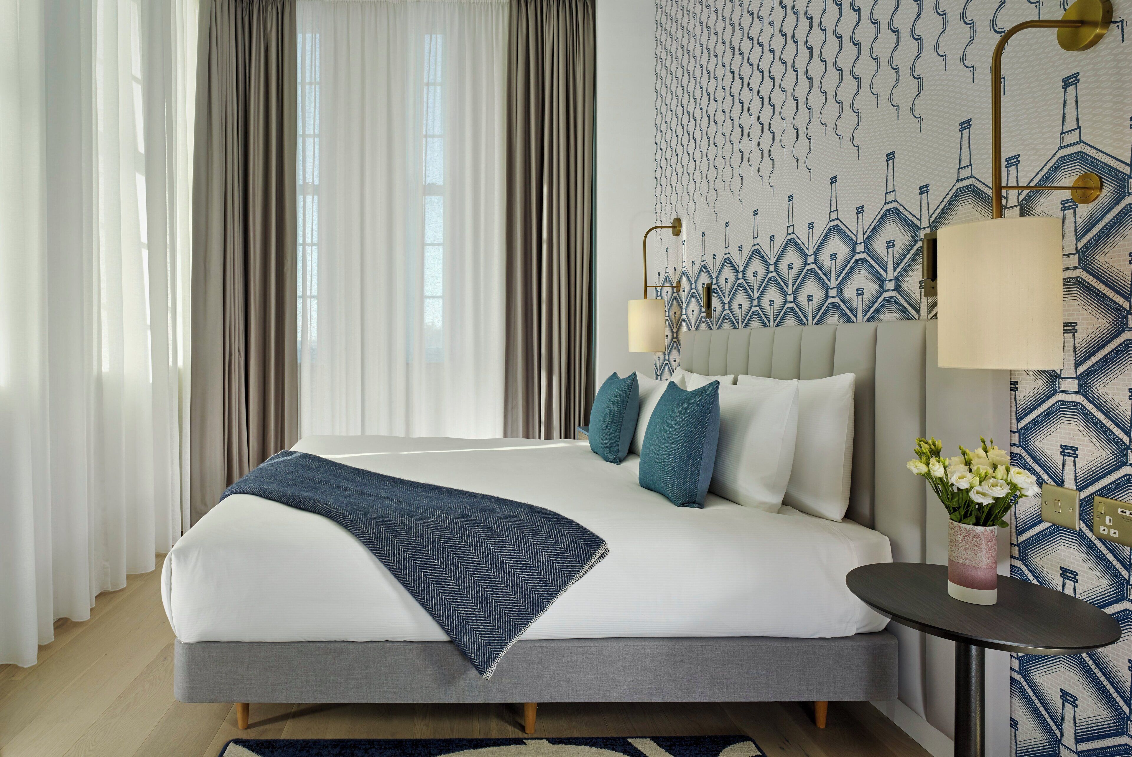 A modern hotel room with a large bed, a bedside table with a vase of flowers, and a patterned wall.