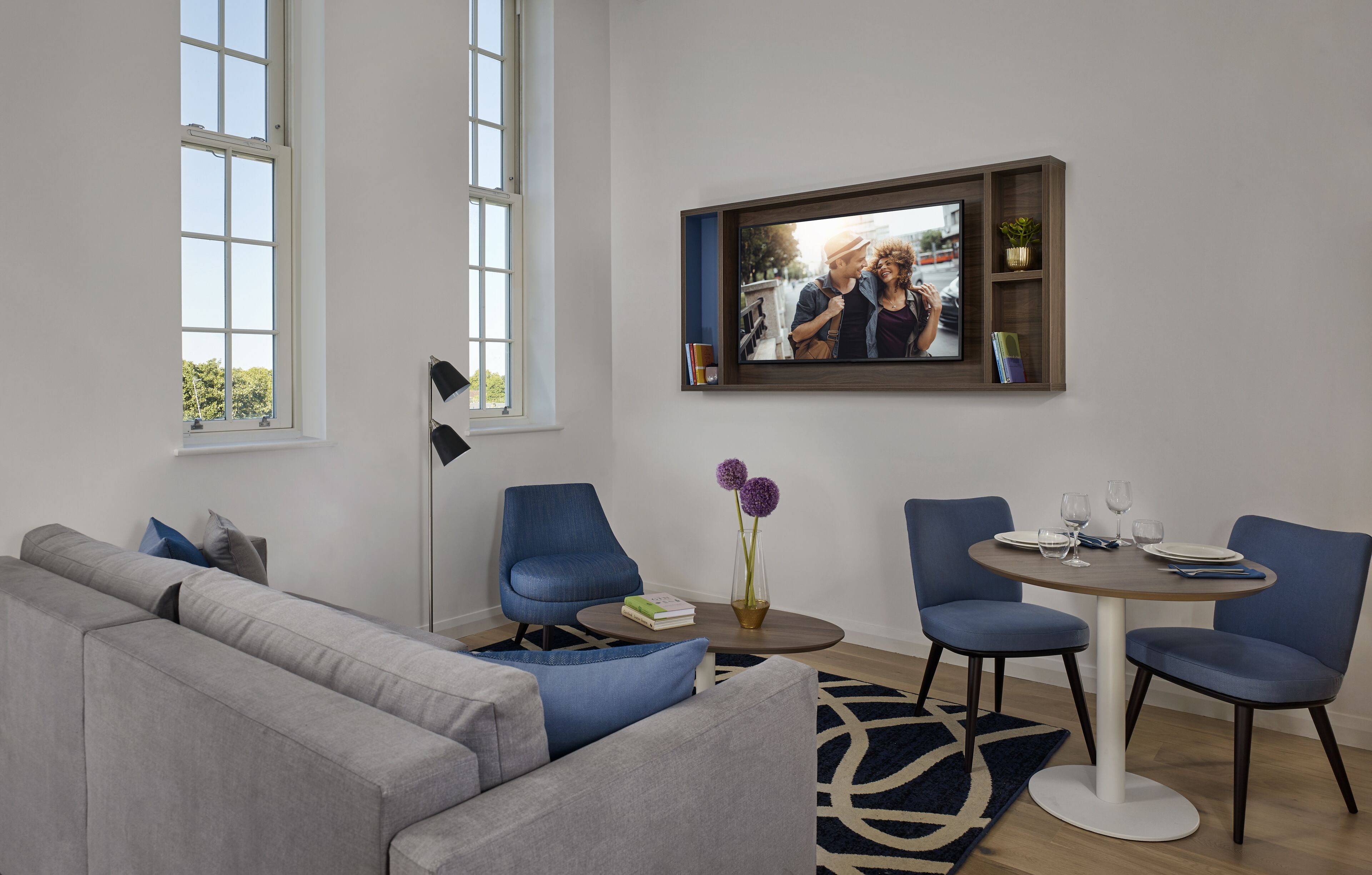 A modern living room with a grey sofa, blue chairs, a round dining table, and a mounted TV.