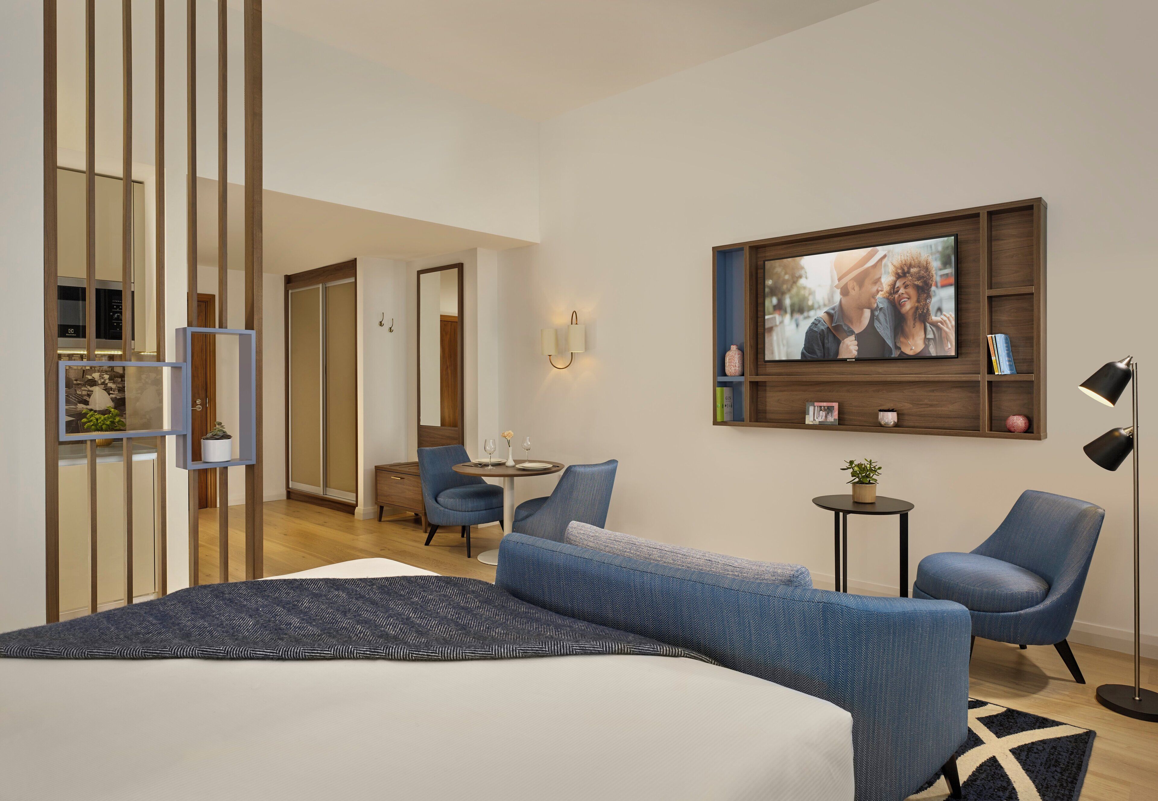 Una habitación de hotel moderna con una cama grande, un área de estar con dos sillones, una mesita pequeña, un televisor montado en la pared y una estantería decorativa con libros y una planta.