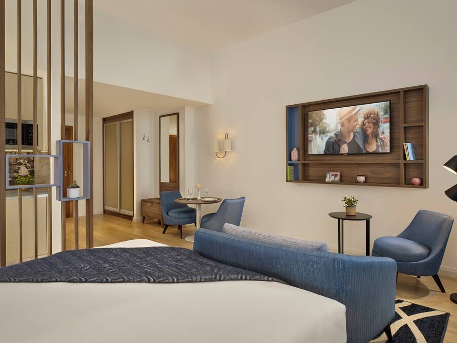Una habitación de hotel moderna con una cama grande, un área de estar con dos sillones, una mesita pequeña, un televisor montado en la pared y una estantería decorativa con libros y una planta.