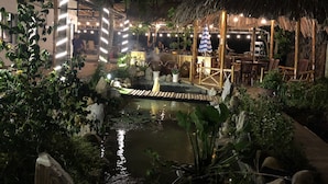 An outdoor dining area with thatched-roof tables and chairs, surrounded by lush greenery and a small pond. - Homestay Songngu Pisces (Can Tho)