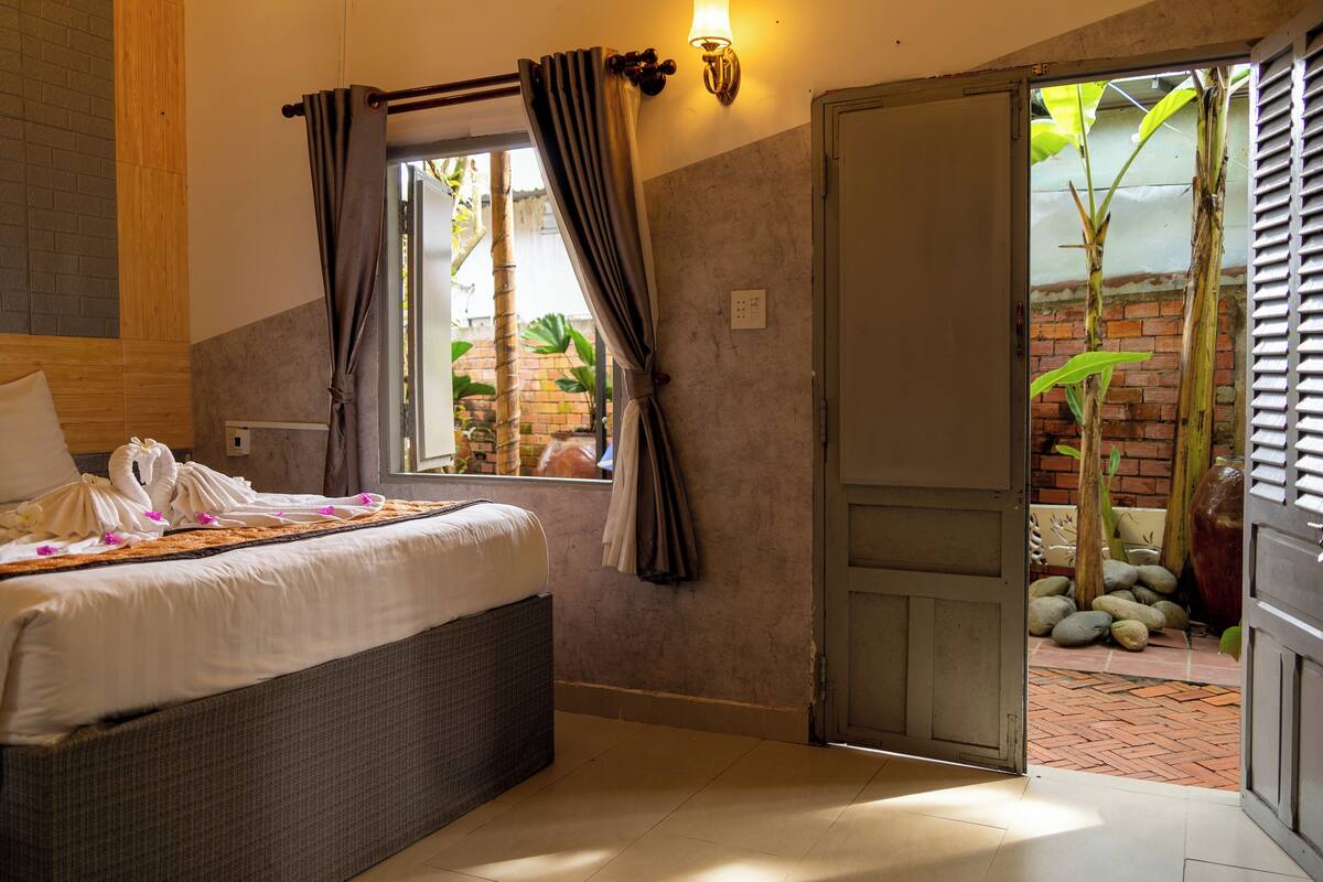 A hotel room with a bed, curtains, a wall-mounted light, and an open door leading to an outdoor area with plants.