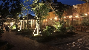 A rustic outdoor seating area with wooden tables and chairs, surrounded by trees and plants, under a clear evening sky. - Homestay Songngu Pisces (Can Tho)