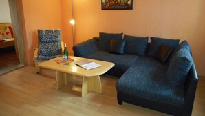 Ein Wohnzimmer mit einem dunkelblauen Sofa, einem Holztisch, einem gemusterten Sessel, einer Stehlampe und einem Stillleben mit Früchten an der Wand.