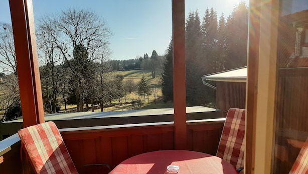 Ein Balkon mit rotem Tisch und karierten Stühlen, der auf eine ländliche Landschaft mit Bäumen und einer Straße blickt.