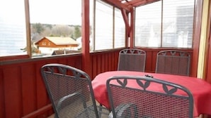 Ein überdachter Außenbereich mit Tisch und zwei Stühlen, mit Blick auf ein Gewässer und ein Gebäude.