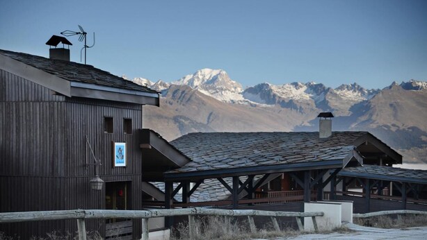 Ein rustikales Gebäude mit einer Holzfassade und einem überdachten Gang, vor einer Kulisse schneebedeckter Berge.