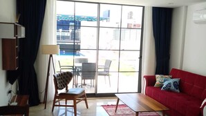 Un salón con un sofá rojo, una mesa de centro de madera, una mesa de comedor con sillas y un amplio ventanal corredero que da a un área exterior.