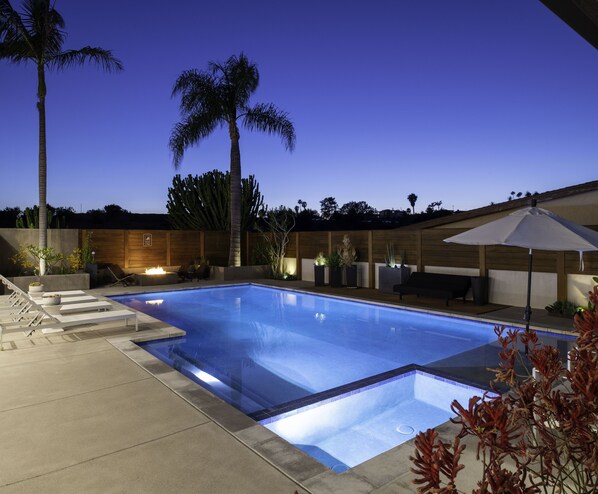 Ein rechteckiger Swimmingpool mit einer gefliesten Kante, umgeben von einer Betonplattform. In der Nähe des Pools befinden sich Liegestühle und eine Holzbank, und ein großer Sonnenschirm spendet Schatten.