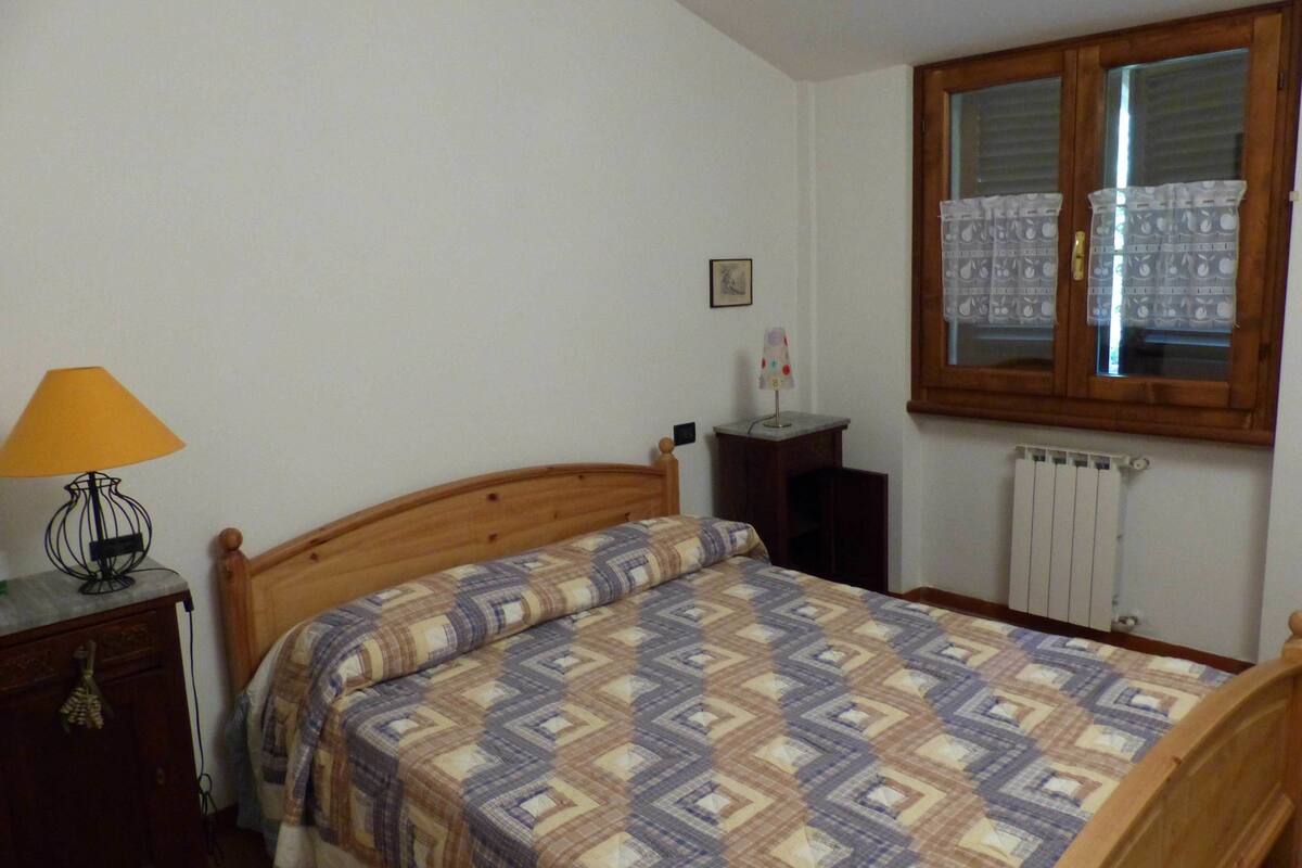A bedroom with a wooden bed, a nightstand with a lamp, a window with curtains, and a radiator.