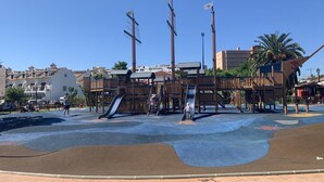 A playground with slides and climbing structures, surrounded by a paved area and buildings in the background. - The Beach House | Family Oasis (Fuengirola Al)