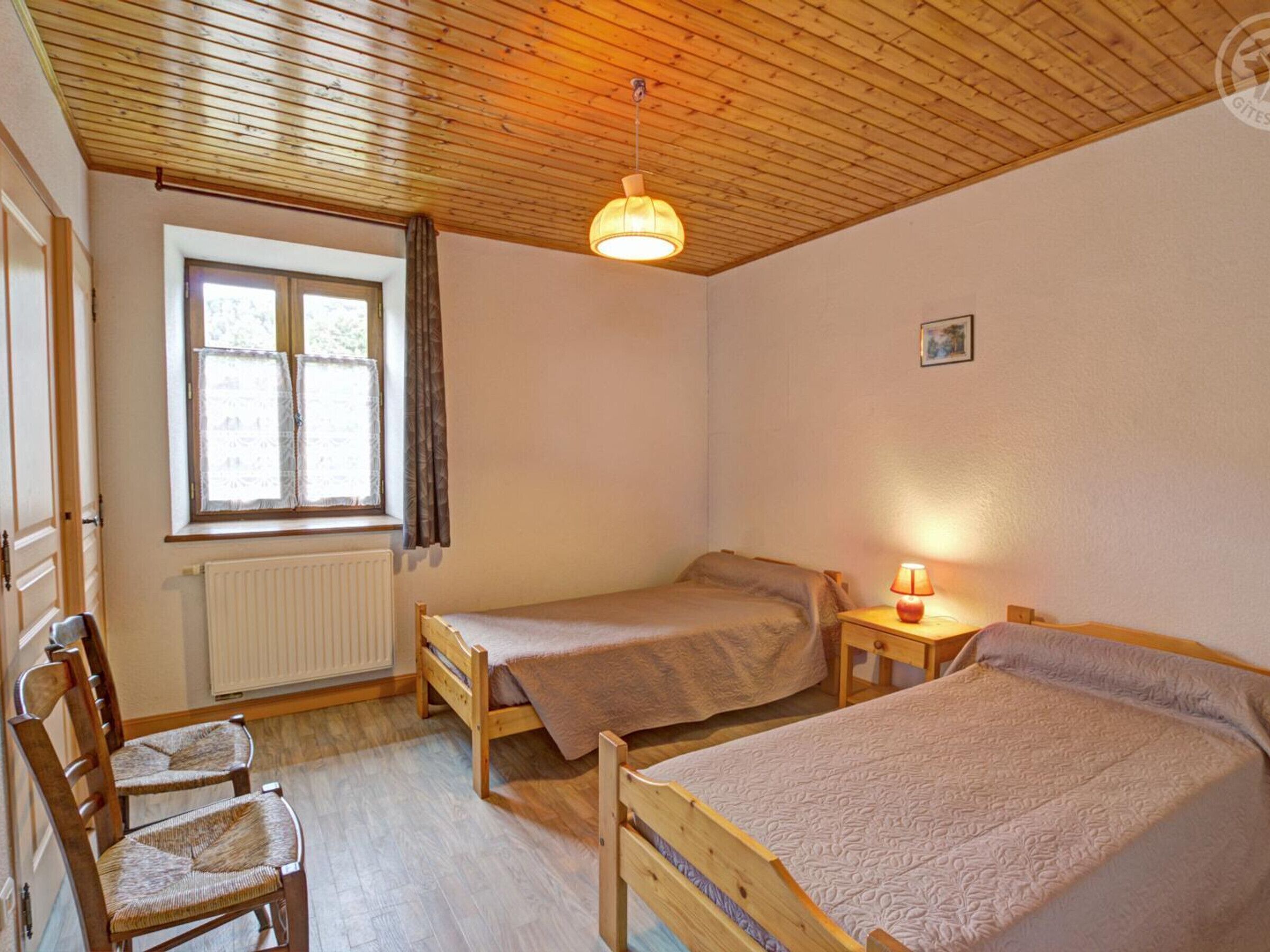 Een kamer met twee houten bedden, een stoel, een raam met gordijnen, een radiator en een hanglamp.