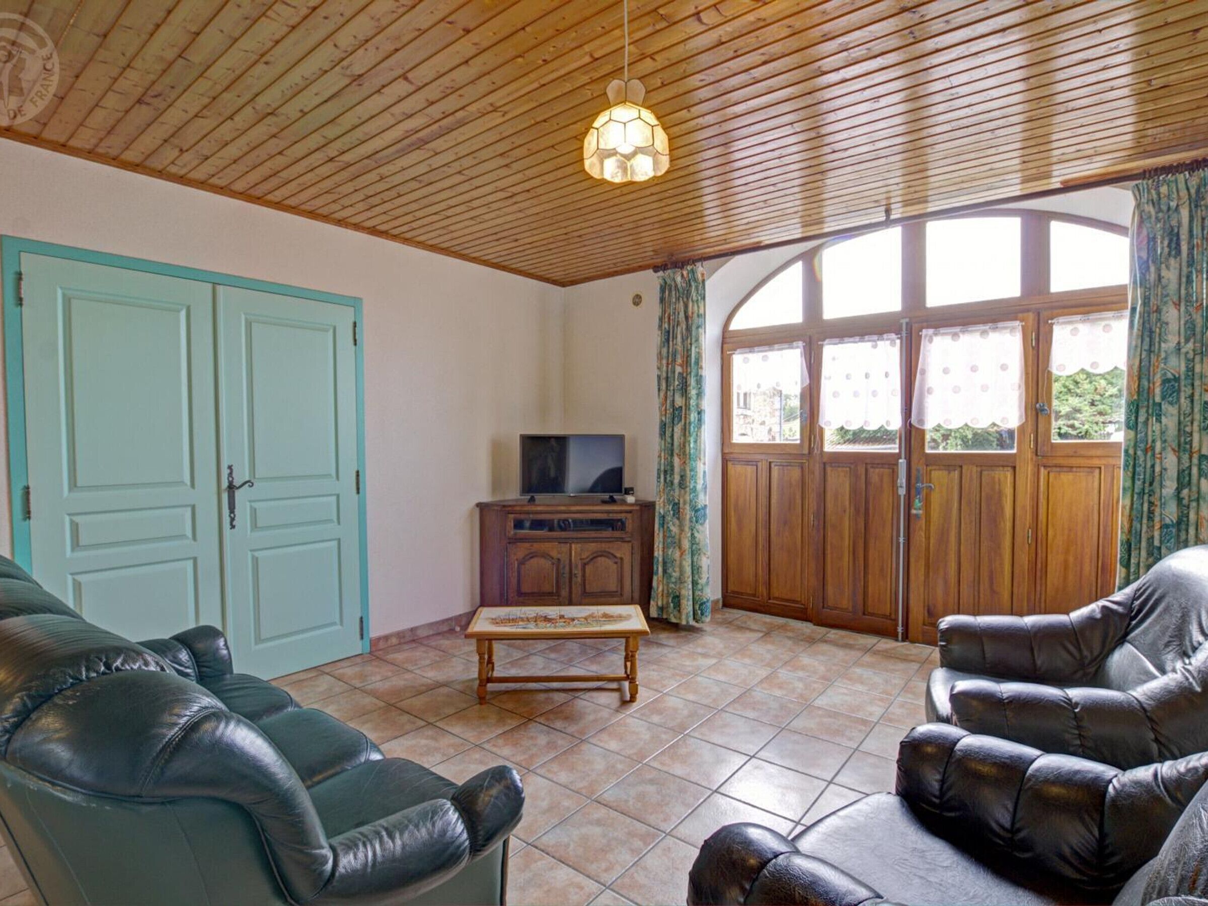 Un salon avec un plafond en bois, un téléviseur posé sur un meuble en bois et une grande fenêtre avec des rideaux.