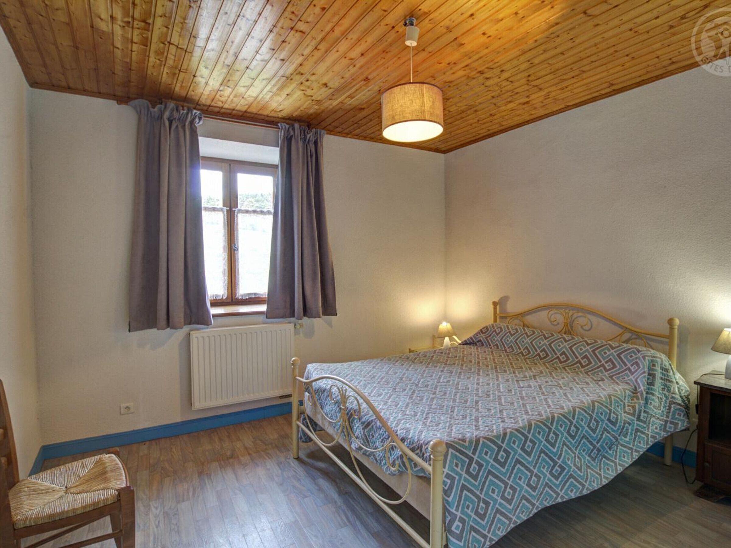Une chambre avec un plafond en bois, un lit recouvert d’une couvre-lit à motifs, une fenêtre avec des rideaux, un radiateur et une chaise.