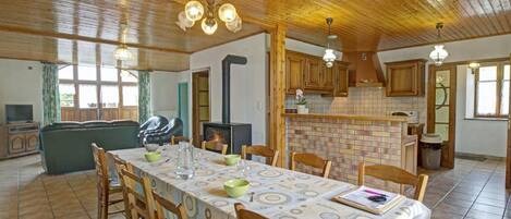 A spacious dining area with a long table set for six, surrounded by wooden chairs. A brick fireplace and wooden cabinets are visible in the background.