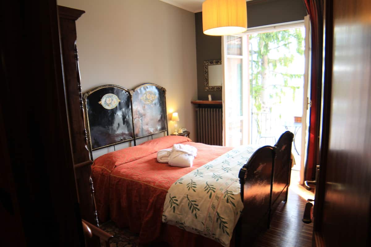 A hotel room with a wooden bed, a red bedspread, a white towel, a nightstand with a lamp, and a window with a view of greenery.