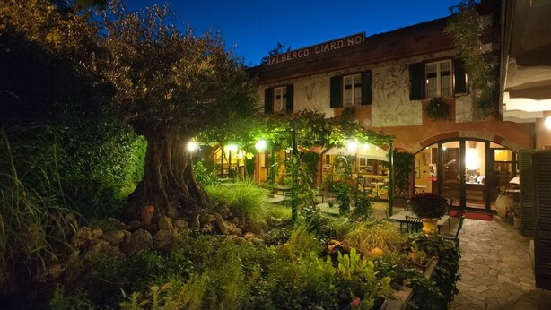 Un edifici amb un rètol que diu "Albergo Giardino" a la nit, envoltat de vegetació exuberant i un camí de pedra.