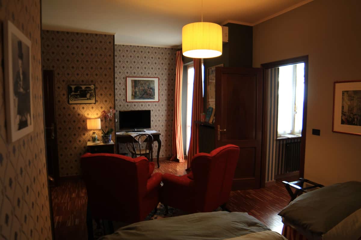 A hotel room with a bed, two red armchairs, a desk with a computer, a lamp, and a large window with curtains.