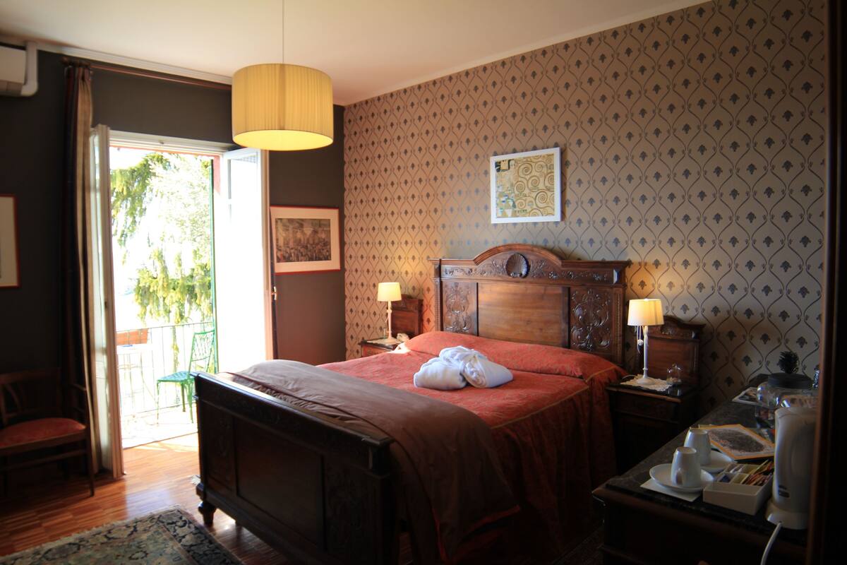 A hotel room with a bed, two bedside lamps, a wooden headboard, a nightstand, a chair, and a view through an open door.