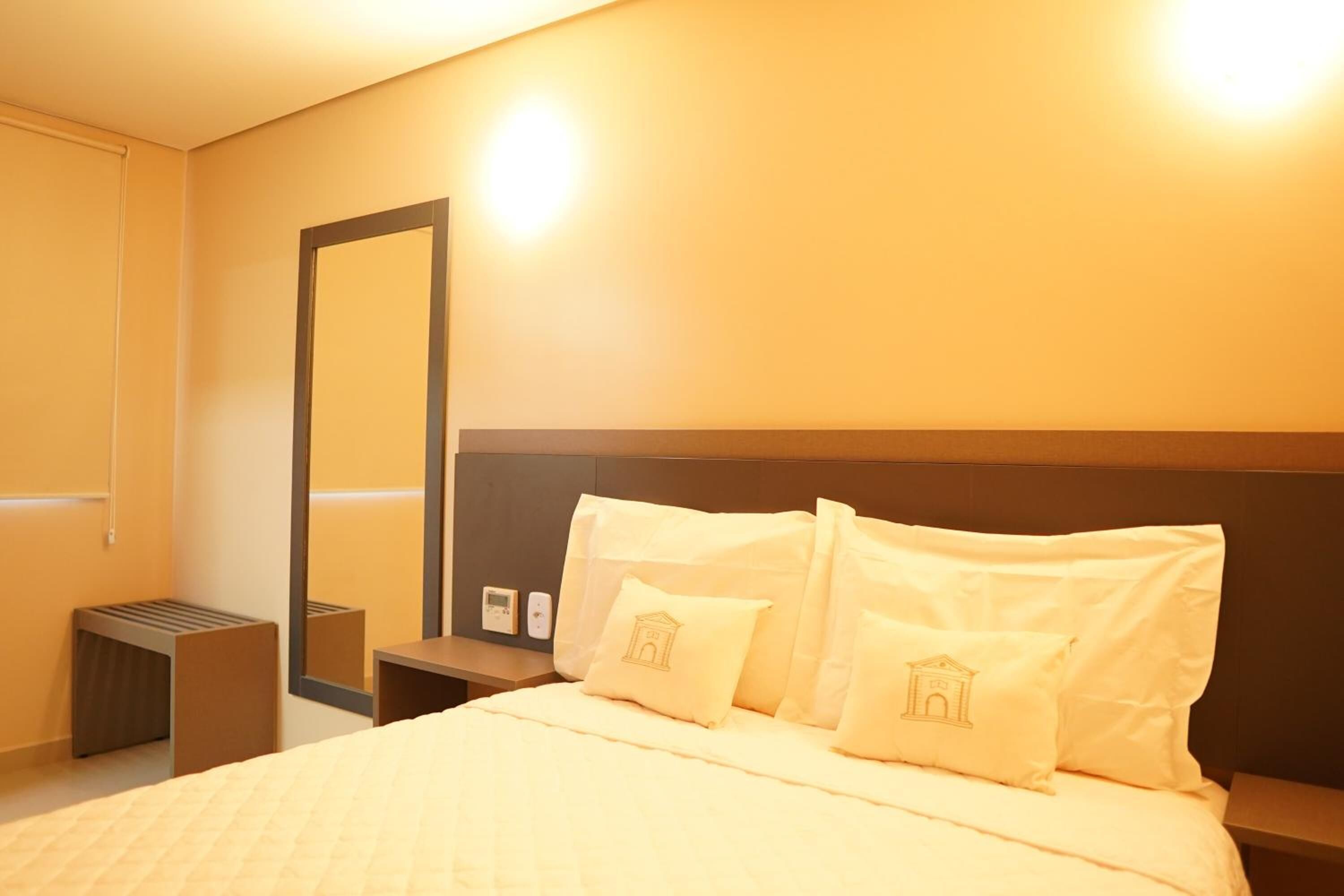 A hotel room with a bed, a nightstand, a mirror, and a wall-mounted thermostat.