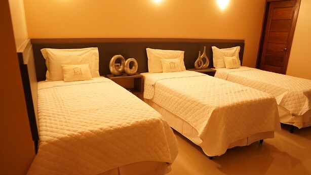 A hotel room with three single beds, each with a white quilted bedspread and matching pillows. The room has a wooden headboard and a nightstand with decorative vases. - Forte Express (Macapa)