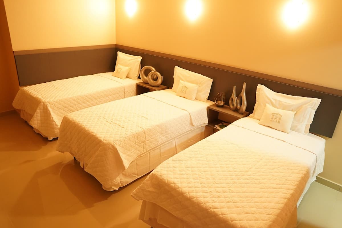 Habitación de hotel con tres camas individuales, cada una con una colcha blanca acolchada y almohadas a juego. Hay mesitas de noche con jarrones decorativos y una lámpara pequeña. La habitación tiene un tono de pared amarillo cálido y un cabecero de madera.