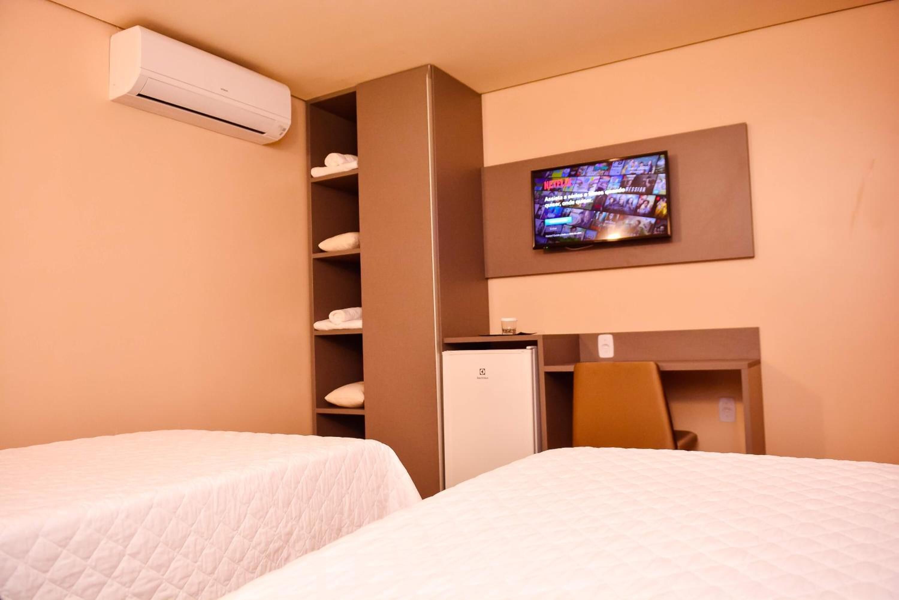 Ein Hotelzimmer mit zwei Betten, einem wandmontierten Fernseher, einer Minibar und einer eingebauten Klimaanlage.
