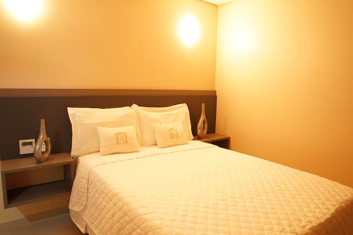 Una cama bien hecha con una colcha blanca acolchada y almohadas a juego, una mesita de noche con un florero, y una lámpara de pared.