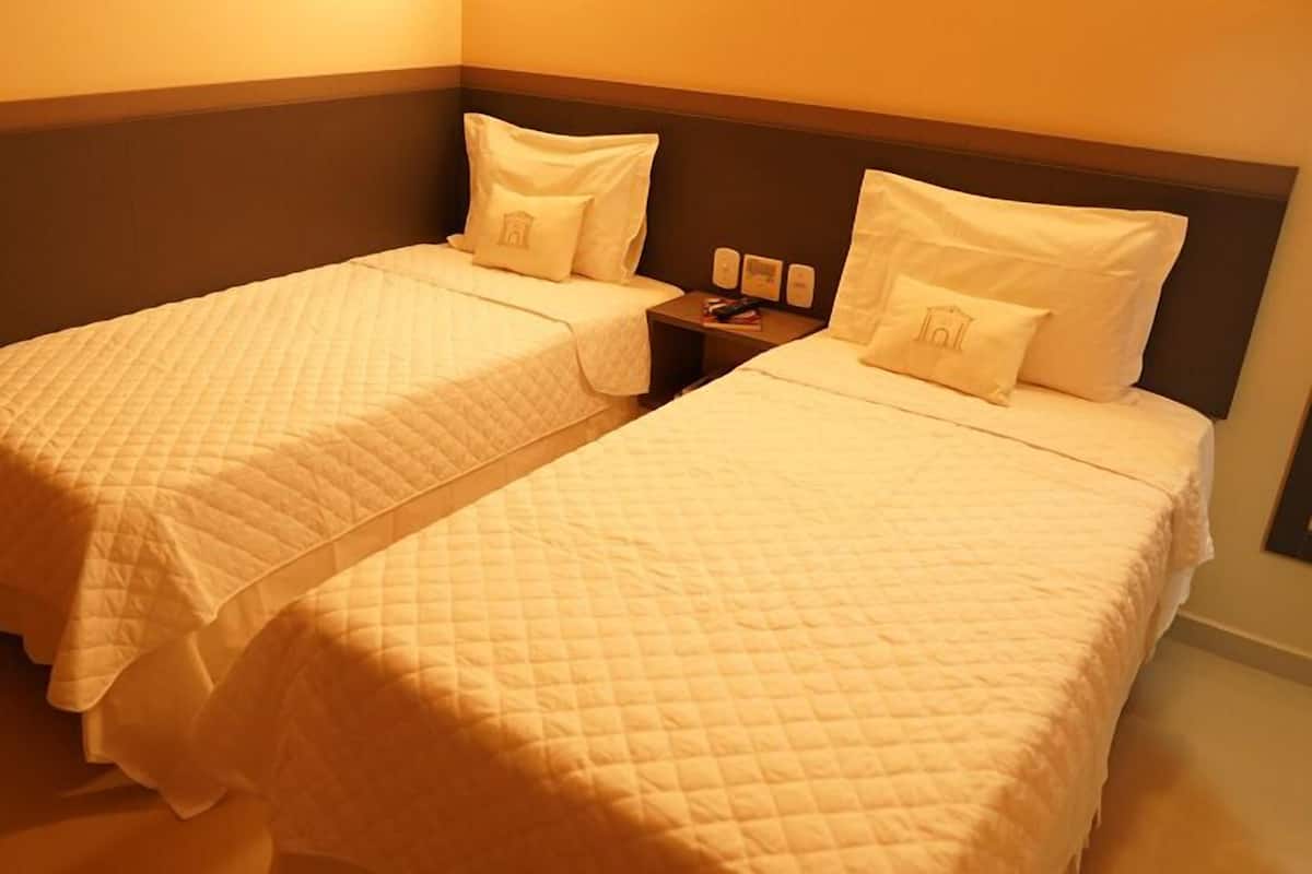 Dos camas juntas con ropa blanca, una mesita de noche con lámpara y un reloj en la pared.