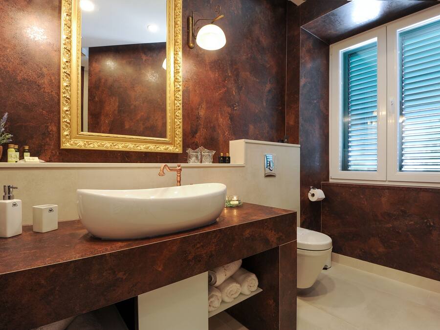 A bathroom with a large oval sink, a tall mirror with a gold frame, a toilet, and a window with blinds.