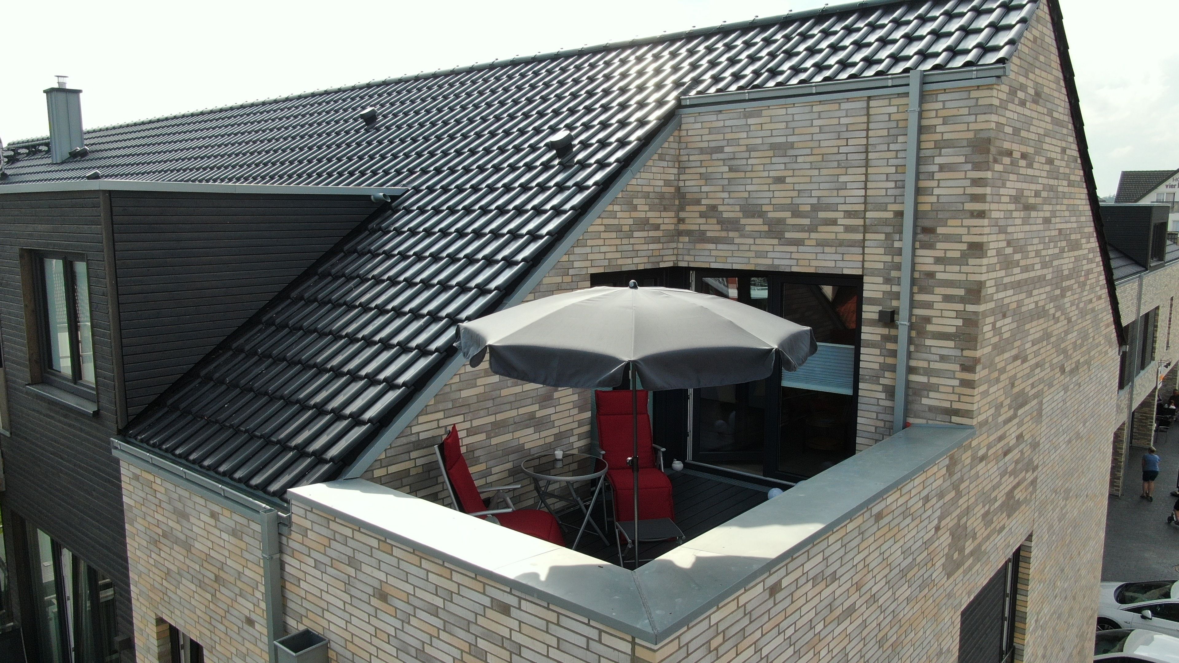 A corner of a building with a sloped roof, a brick wall, and a small outdoor seating area with a table, chairs, and an umbrella.