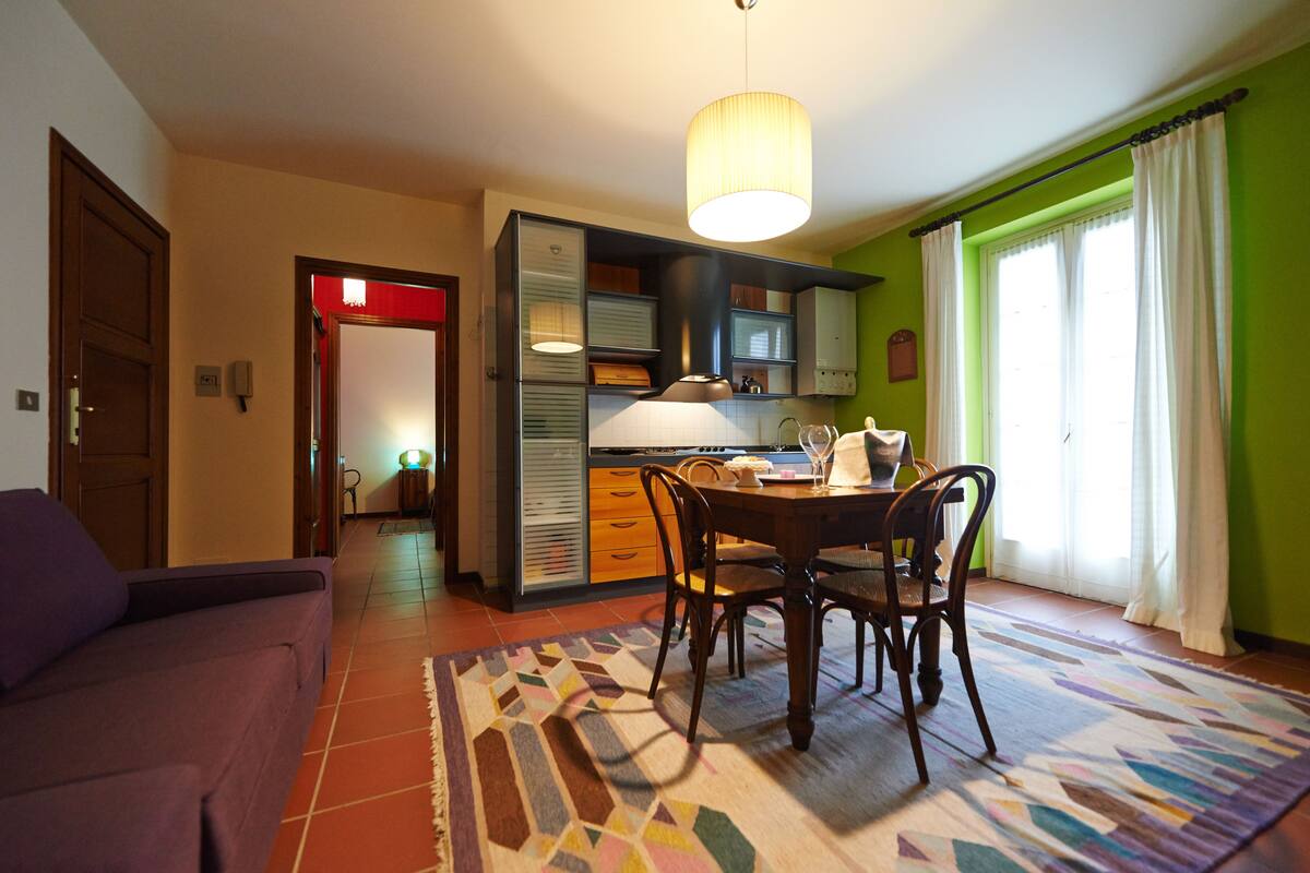 A dining area with a table and chairs, a hanging light fixture, and a view of a green wall and window with curtains.