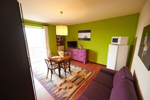 Un espacio habitable compacto con comedor, un sofá morado, una cocina con microondas y un televisor.
