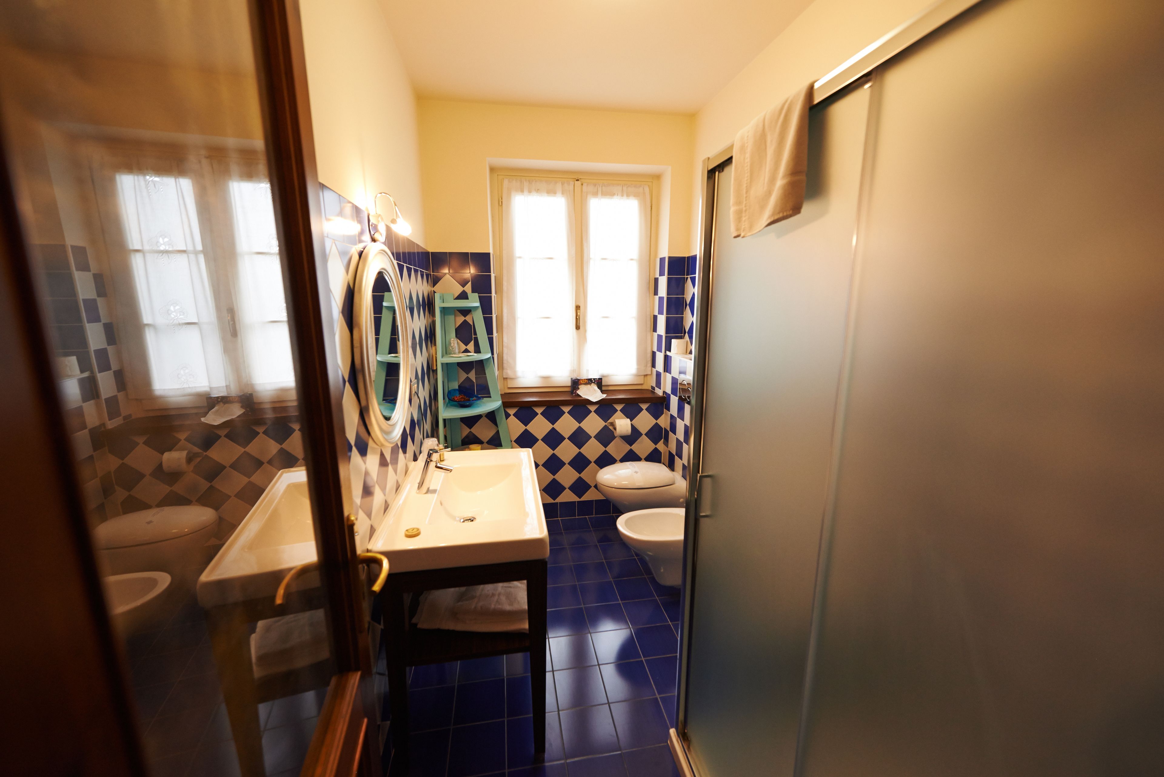 A bathroom with a sink, toilet, and bidet. There is a window with curtains and a towel hanging on the door.