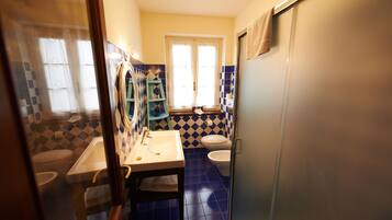 Un baño con lavamanos, inodoro y bidé. Hay una ventana con cortinas y una toalla colgada en la puerta.