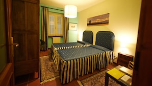 A room with two beds, a wooden wardrobe, a window with curtains, a painting on the wall, and a table with a lamp. - Felicin - Dimora del Centro (Monforte d'Alba)