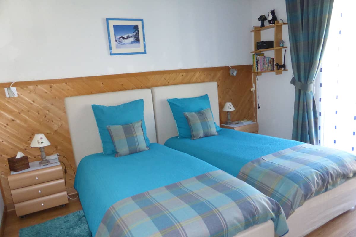 A double bed with blue bedding and plaid throw pillows, a bedside table with a lamp, a small shelf with books, and a framed picture on the wall.