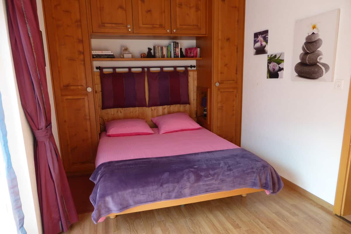 A wooden bed with pink and purple bedding, wooden cabinets above, and a small shelf with books and decorative items.
