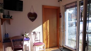 A room with a wooden floor, a white chair, a table with a purple tablecloth, a heart-shaped mirror on the wall, and a sliding glass door leading outside. - B&B Les Acacias (Bagnes)