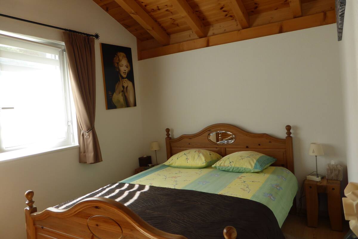 A bedroom with a wooden ceiling, a bed with a headboard, a window with curtains, and a wall-mounted picture.