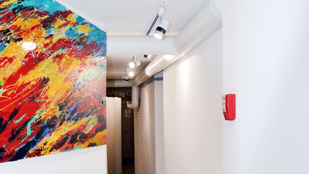 A narrow hallway with a colorful abstract painting on the left wall, a white ceiling with recessed lighting, and a red fire alarm on the right wall. - Hostel Train Station (Kyiv)
